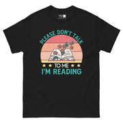 A retro-style design with a semi-circle background in shades of red, orange, and yellow. An open book with flowers growing from it is in the foreground. The teal text "PLEASE DON'T TALK" curves above the book, and "TO ME I'M READING" is below it, with yellow stars separating "TO ME"